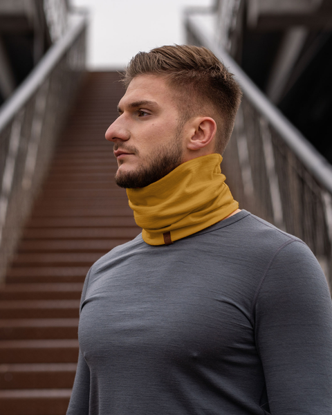A man with short light brown hair and a beard is shown in profile, looking to the left. He is wearing a spicy yellow menique neck gaiter and a gray long-sleeved shirt, standing on concrete stairs with metal railings.