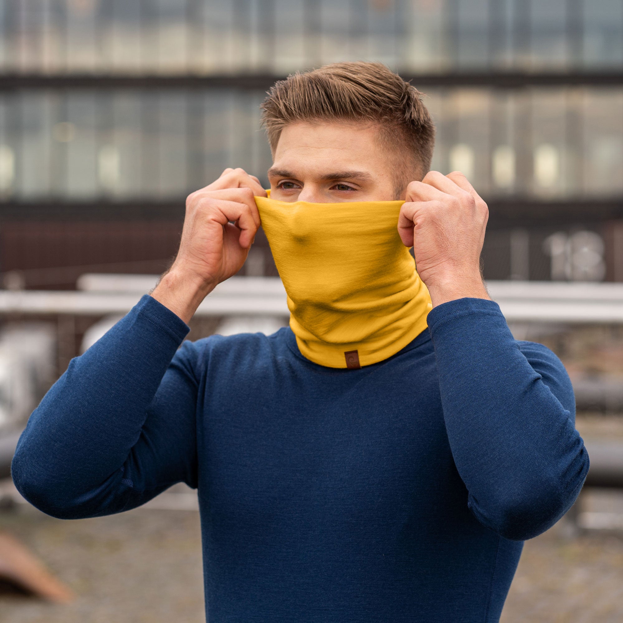 A person with light eyes is wearing a spicy yellow menique beanie that is pulled down to cover part of their forehead and a matching neck gaiter that is pulled up to cover their mouth and nose. They are wearing a dark long-sleeved shirt, and the blurred background suggests an outdoor setting with trees and foliage in muted tones.