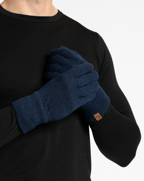 A person wearing blue knit gloves, possibly merino wool, is shown in a close-up studio shot against a white background. The gloves have ribbed cuffs and a small brown tag is visible on the wrist of the right glove. The person is wearing a long-sleeved black shirt.