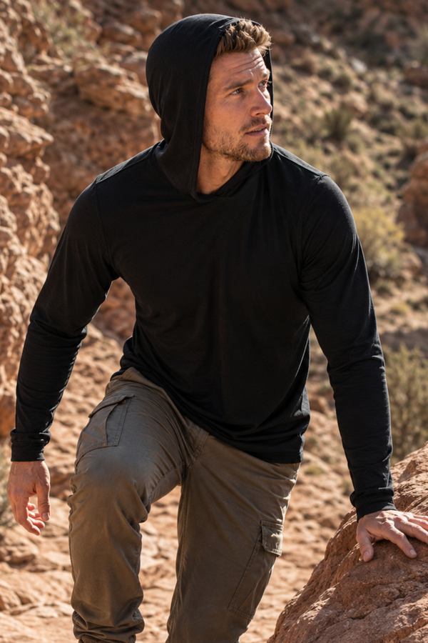 An outdoor shot of a man hiking along a rugged, sunlit rocky trail. He is looking off to the left, wearing a black long-sleeved hooded shirt with the hood down and olive green cargo pants.