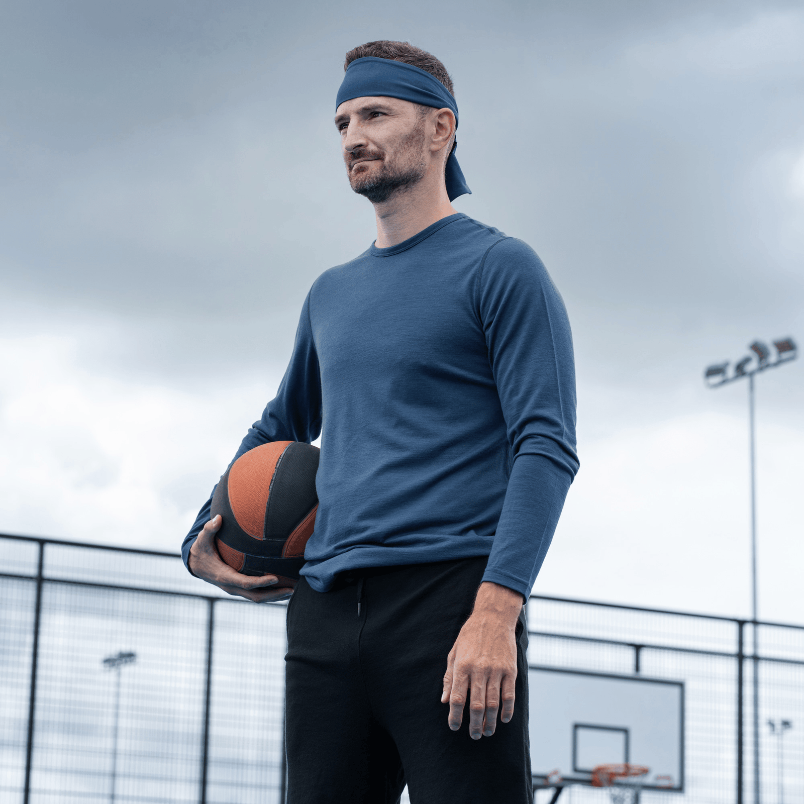 A slightly closer shot of man with a blue headband and dark blue long-sleeved shirt smiling on an outdoor basketball court while holding a basketball.