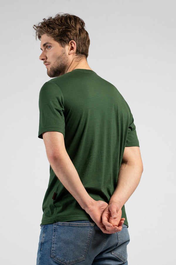 A man stands with his back partially to the camera, glancing back over his left shoulder with his hands clasped behind him. He is wearing a dark green short-sleeved t-shirt and blue jeans.