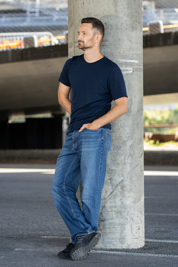 A smiling man stands outdoors with his right hand in his pocket, looking toward the right. He is wearing a navy blue short-sleeve crew neck t-shirt and light beige shorts. He is positioned in front of a textured, rusted metal barrier.