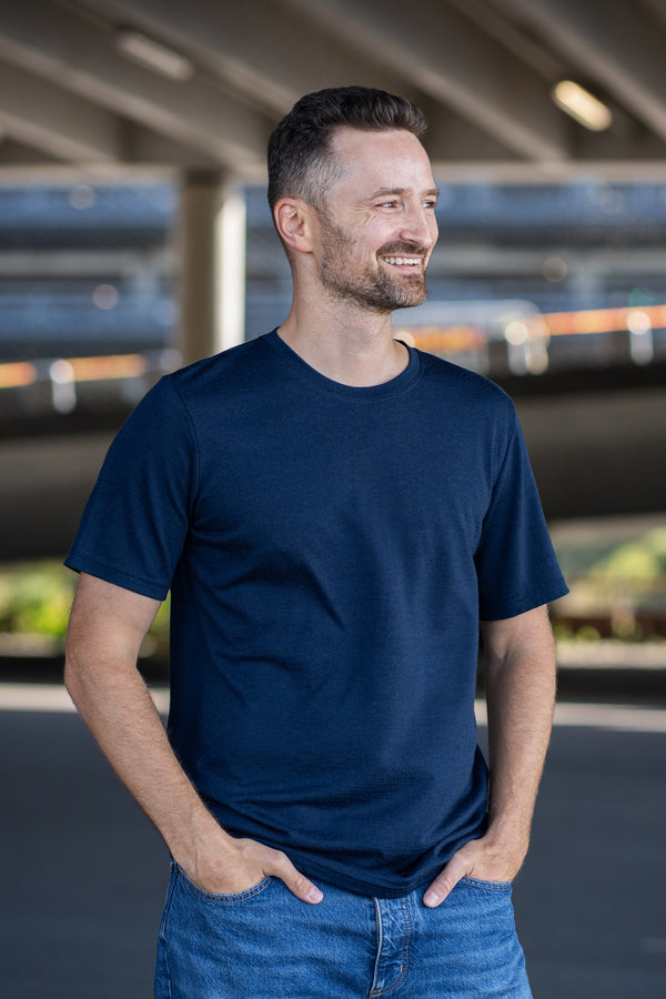 A man with a short beard smiles and looks to the right while standing with both hands tucked into the front pockets of his blue jeans. He is wearing a navy blue short-sleeve crew neck t-shirt. The background is a shaded outdoor area beneath a concrete overpass.