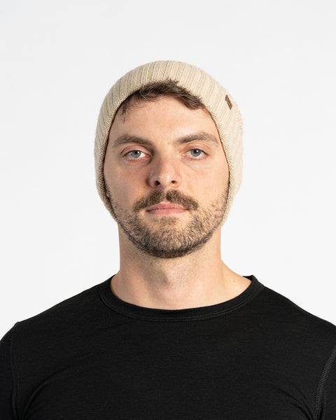 A person with light skin, short brown hair, a mustache, and light-colored eyes is shown in a headshot against a white background. They are wearing a creamy beige knit beanie with a small brown tag on the side and a black long-sleeved shirt. They are looking directly at the camera with a neutral expression.
