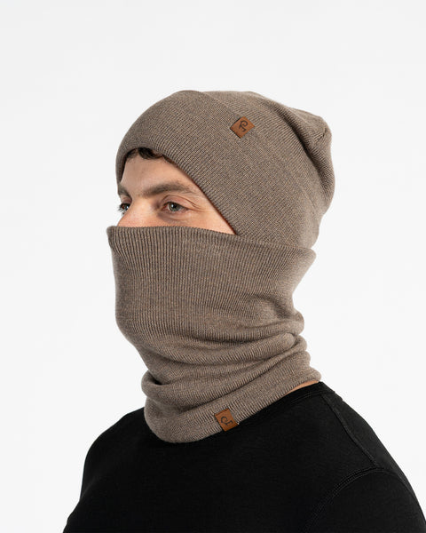 A male model, viewed in profile, wears a  brown beanie and a matching neck gaiter that covers his nose and mouth. Both the beanie and gaiter feature a small, brown leather patch with a Menique logo. He has a short beard and dark hair. The background is solid white.