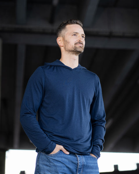 A man standing outdoors under a concrete structure, looking off-camera with a neutral expression. He is wearing a navy blue lightweight hoodie and blue jeans, with his hands tucked into his front pockets.