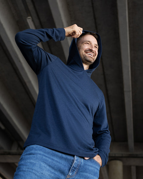 A smiling man standing outdoors under a concrete overpass. He is wearing a lightweight navy blue hoodie and blue jeans, and is pulling the hood over his head with one hand while looking off-camera.