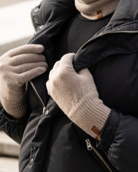 A person wearing menique merino wool knit gloves and a matching neck gaiter is shown adjusting the zipper of a black puffer jacket outdoors. The gloves have ribbed cuffs and small brown tags. The background shows a blurred urban environment with concrete steps or seating.
