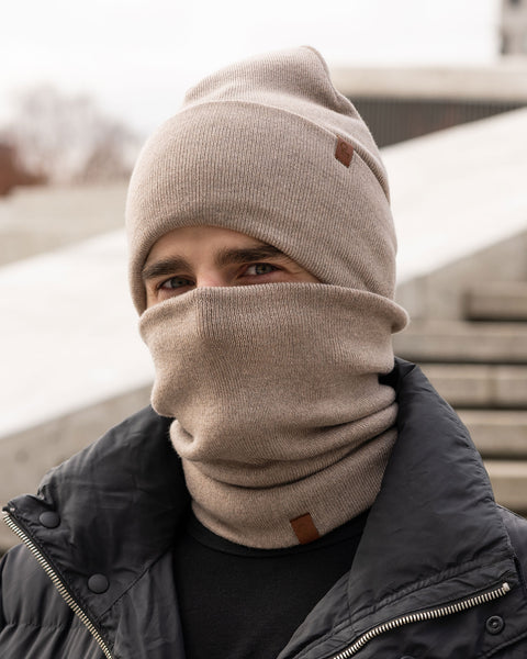 A person with light brown hair and eyes is shown outdoors, wearing a creamy beige knit beanie pulled down to their eyebrows and a matching neck gaiter covering their mouth and nose. They are wearing a black puffer jacket. The beanie has a small brown tag on the side. The background is blurred, suggesting an urban environment with steps or concrete structures. Only the person&#39;s eyes are visible above the neck gaiter.