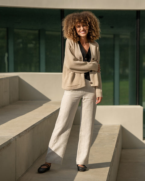 Woman in a beige knit sweater, black linen shirt and natural linen pants standing in front of a modern building