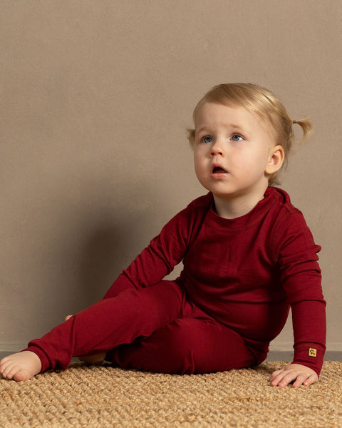 a girl sitting on the floor, wearing the Kids&#39; Long Sleeve Set 160gsm Merino Wool thermal set in red color
