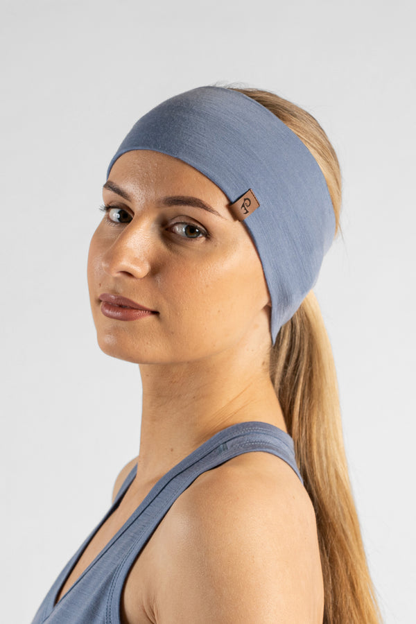 A studio portrait of a blonde woman looking directly at the camera with her body angled slightly away. She is wearing a dusty blue headband over her pulled-back hair, featuring a small brown leather logo patch. She is wearing a matching dusty blue sleeveless top against a plain light grey background.