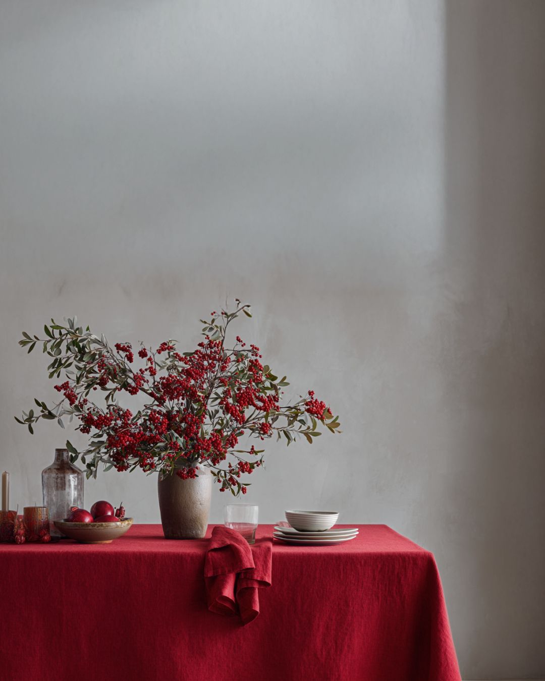 Table setting with a vase of red flowers on a red tablecloth against a plain wall.
