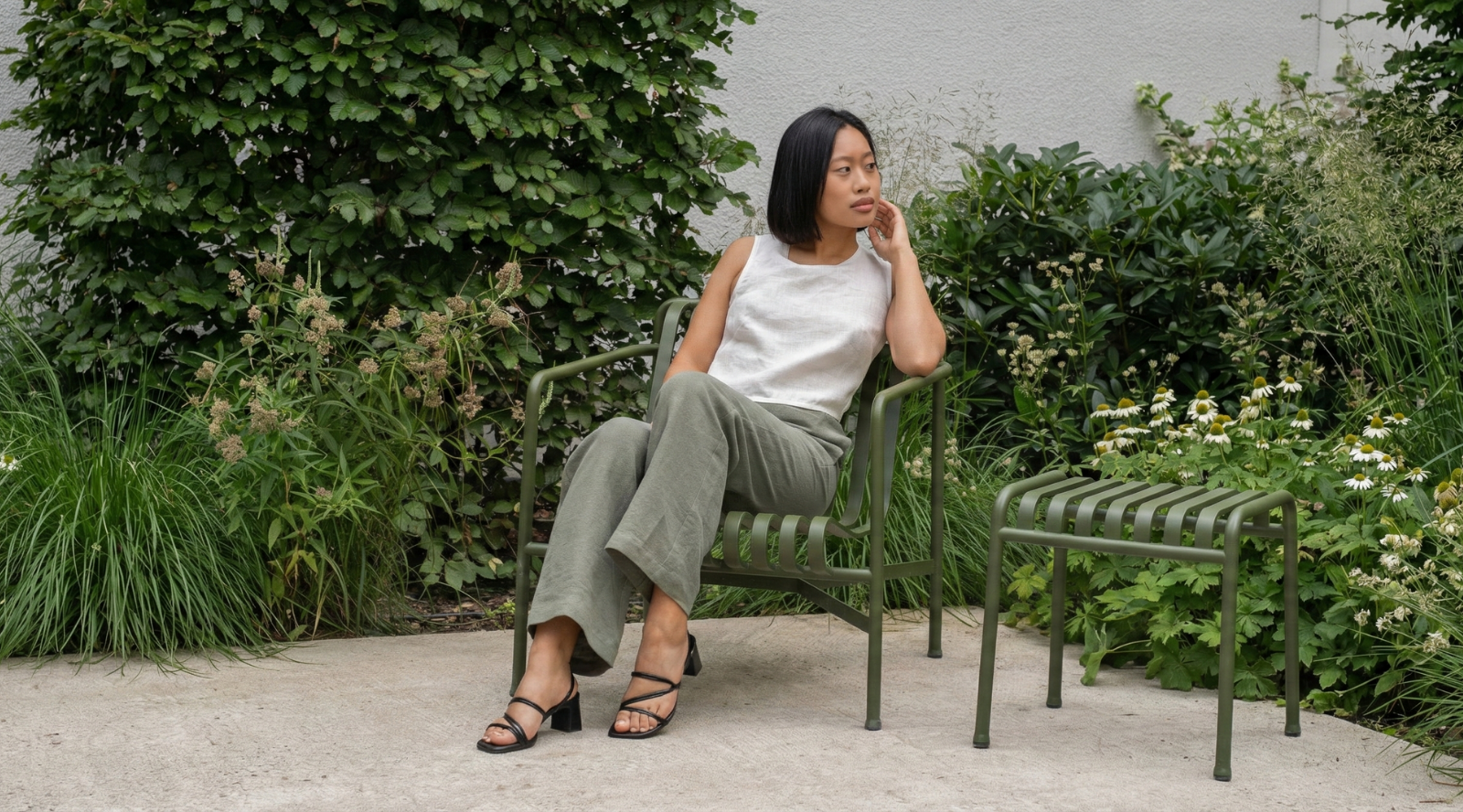 A woman with short black hair is sitting on a green metal chair in a garden, wearing a sleeveless white linen top and relaxed-fit olive green linen trousers. She is looking to the side with a calm expression, surrounded by lush greenery and plants, creating a peaceful outdoor setting.