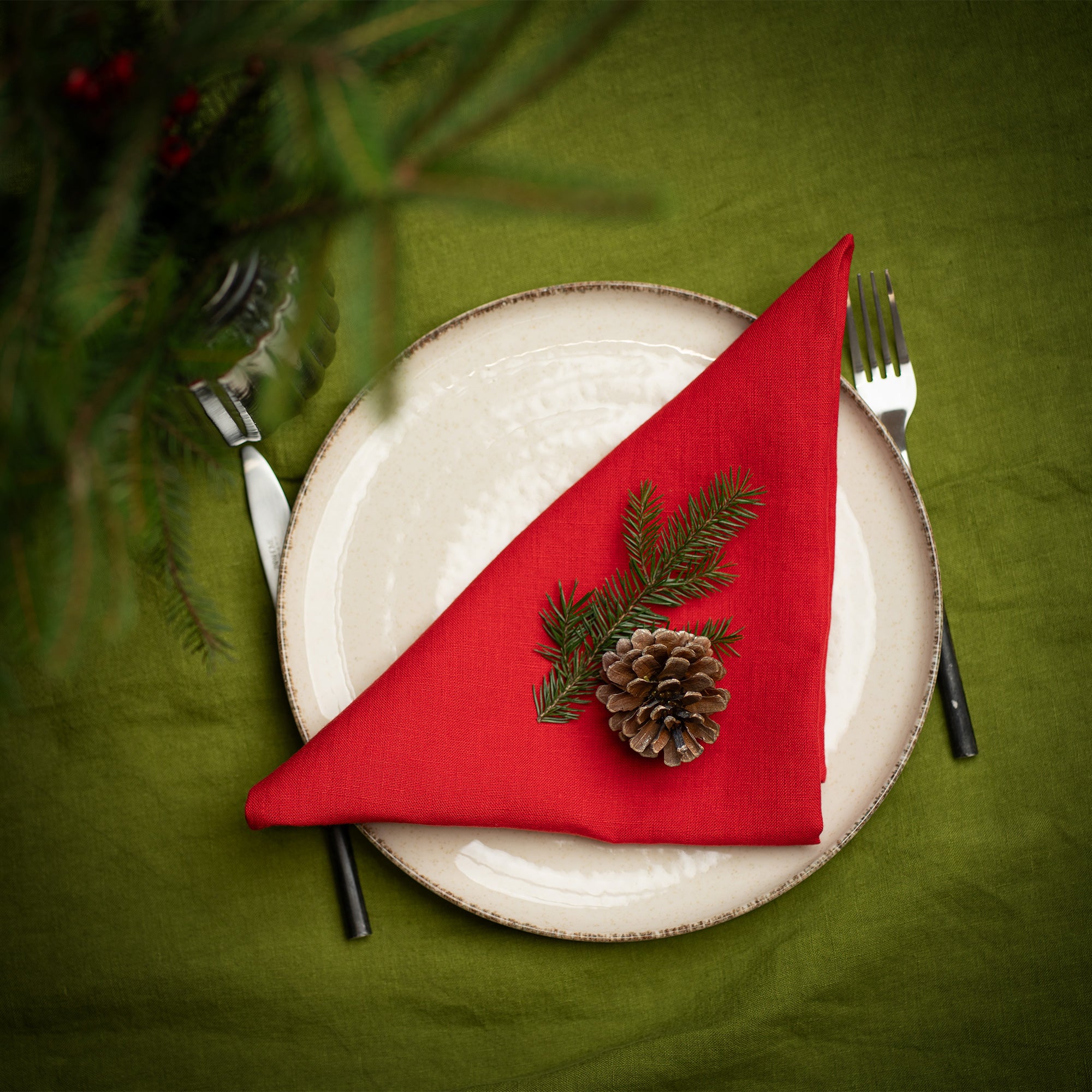 A top-down view of a Christmas place setting with a cream-colored plate on a green tablecloth. A red linen napkin is folded into a triangle and decorated with a pinecone and fir sprig. Silverware is placed on either side, and evergreen branches appear at the top edge of the image.