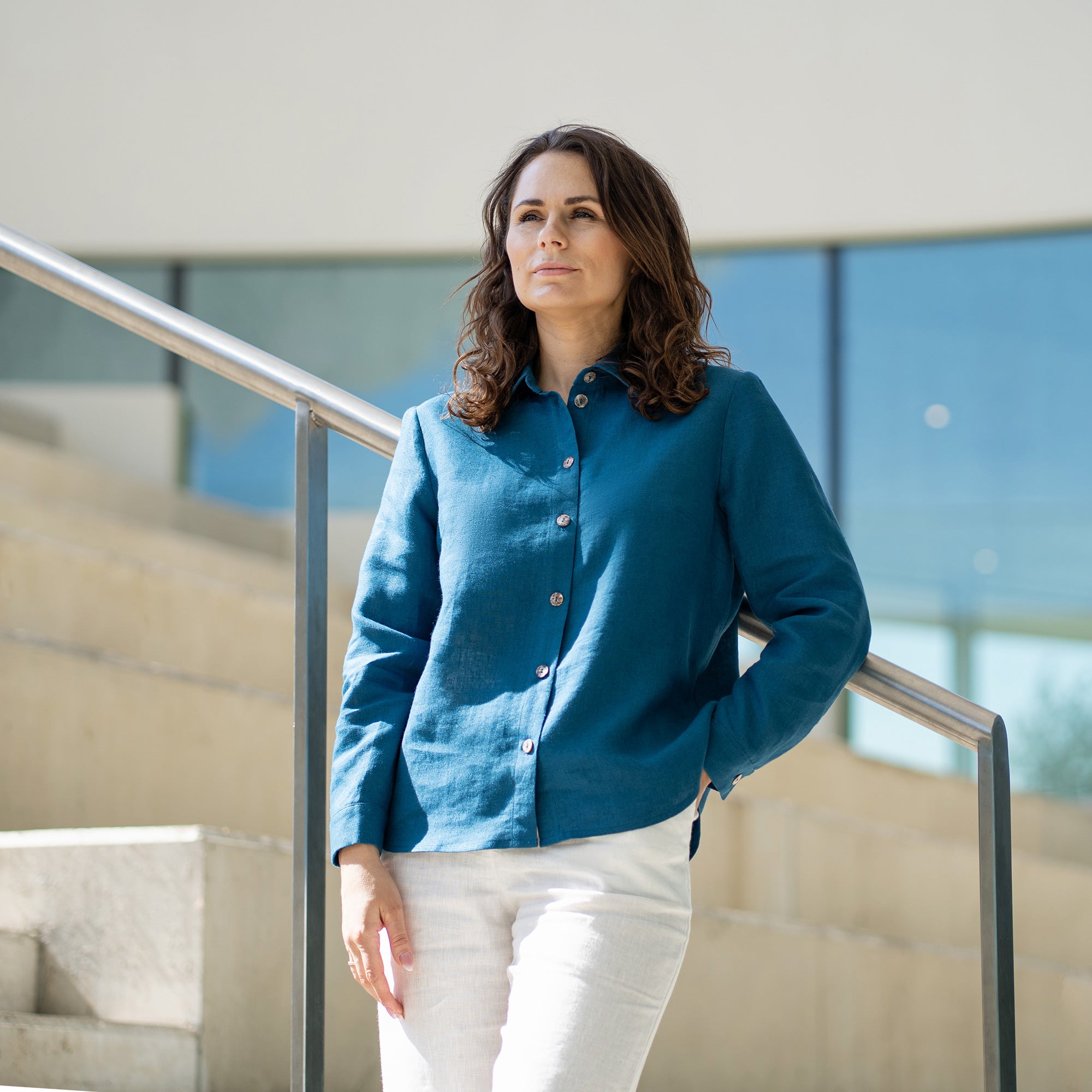 A woman with brown hair, wearing a light blue button-front shirt and wide-leg white trousers, stands outdoors against a modern concrete structure.
