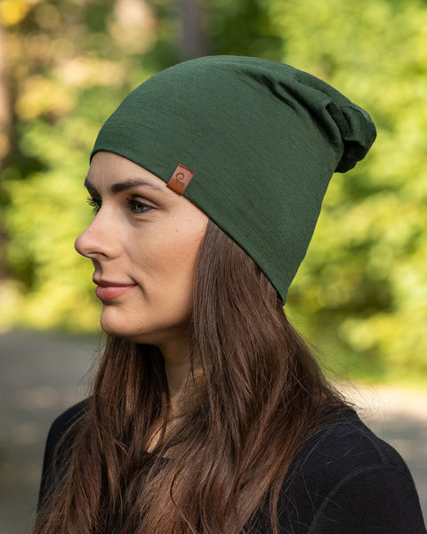 The picture features a side profile head-shot of a woman wearing the dark green unisex beanie. It is made from merino wool, has a small logo on the side at the front. Behind her is a blurred view of trees .