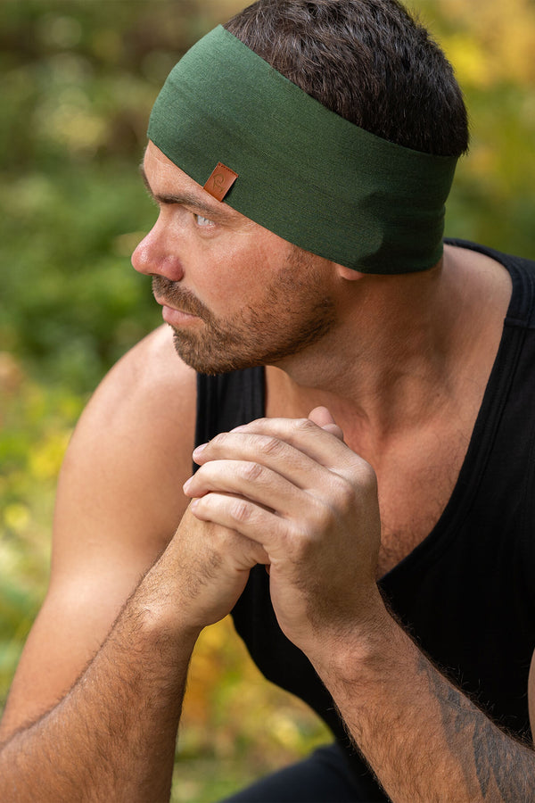 The picture features a man sitting down in a forest. He is looking to the side, with his hands on his knees, clasped together. He is wearing a dark green menique merino headbands with a small logo on the side at the front. He is also dressed in a sleeveless menique merino black top.