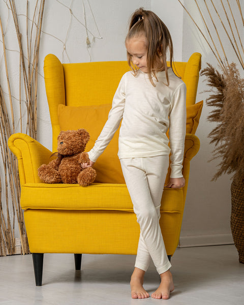 A girl standing by a yellow armchair, holding a teddy bear by the hand, wearing the menique Kids&#39; 160 Merino Pants in Natural color.
