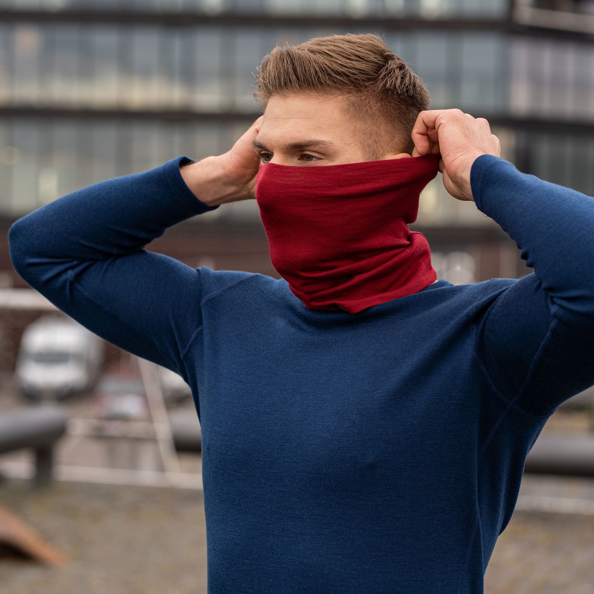 A man, standing outside in a forest during fall season, wearing the Menique Men's Merino Beanie and neck Gaiter Set in the color Royal Cherry.