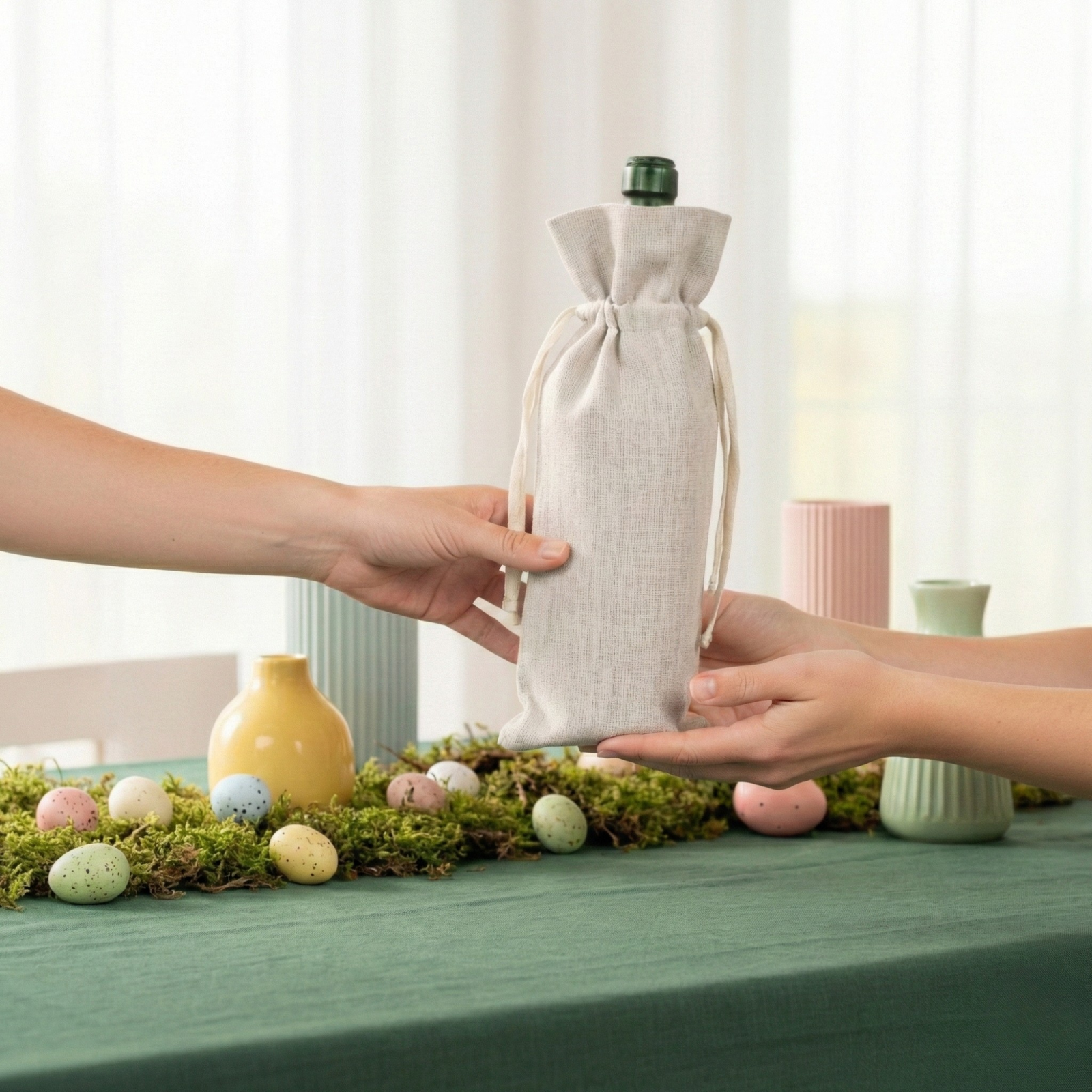Three linen wine bottle gift bags in red, green, and natural tones displayed on a spring table with flowers, candles, and pastel décor.