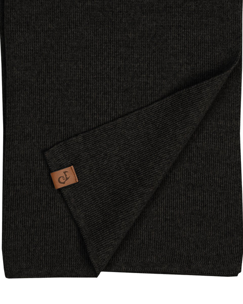 A close-up flat lay of a black knitted merino wool scarf. The bottom edge is folded up diagonally to reveal a small, rectangular brown leather logo patch sewn near the hem. The texture of the knit is clearly visible. The background is pure white.