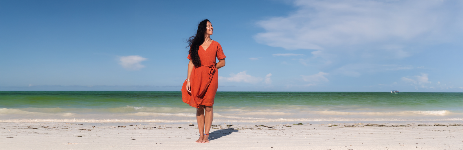 A woman with long dark hair stands on a sandy beach, facing away from the camera. She wears a long-sleeved orange dress and is barefoot. The ocean is in the background with light waves, and the sky is blue with a few clouds.