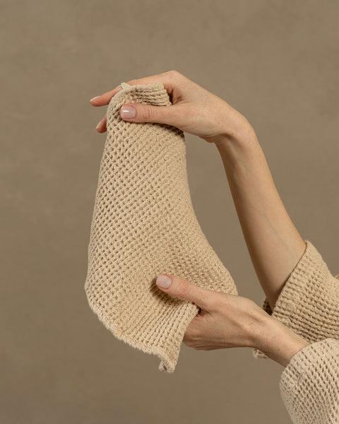 A woman holding a Waffle Washcloth Face Towel of 30x30cm dimensions in her hands. The towel is in sand brown color, as well as the visible sleeves of the robe she is wearing, that is made from the same fabric.