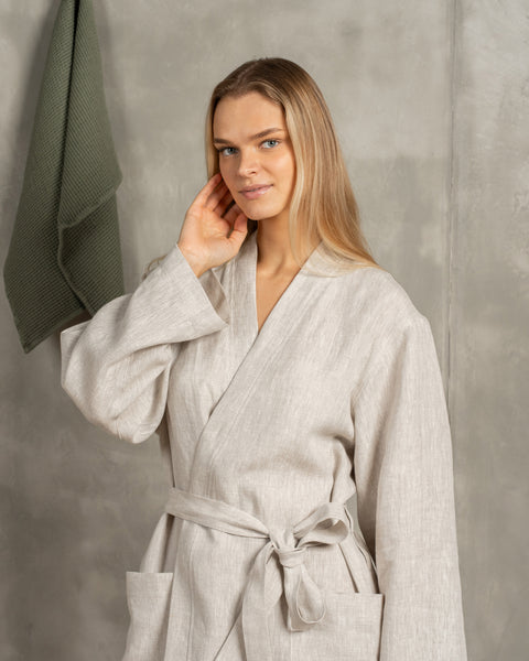 A woman with blonde hair wearing a natural color 100% linen menique robe with a tied belt, touching her face, against a textured grey background.