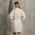 The menique women's linen bathrobe in natural color. The woman is standing in front of a gray wall with a wooden hanger on it. There is a linen waffle towel on the hanger. She has in the pocket, the other put lightly on her stomach.