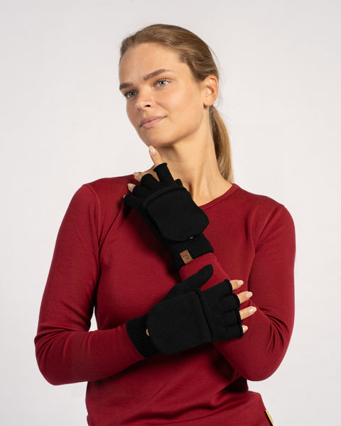 Frontal portrait of a woman wearing black convertible mittens, one mitten flap folded back to use as fingerless gloves while she rests a hand near her chin.