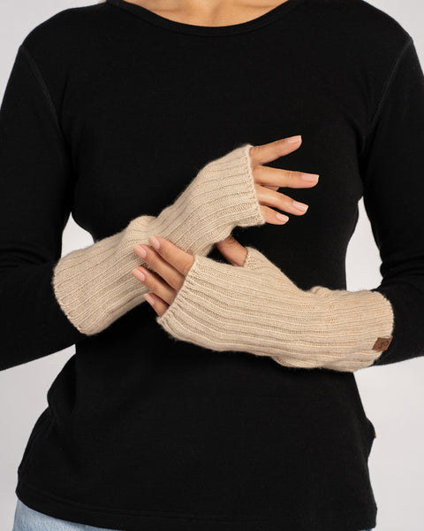 Close-up of a model’s crossed hands wearing creamy-beige ribbed wrist-length merino &amp; cashmere fingerless gloves against a black top.