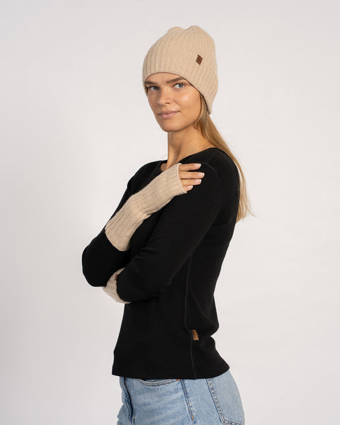 Three-quarter portrait of the model wearing creamy-beige wrist gloves and a matching beanie, arms crossed and smiling slightly.
