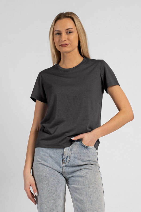 A studio portrait of a blonde woman standing and looking directly at the camera, with her left hand casually tucked into the front pocket of her light-wash blue jeans. She is wearing a regular fit, charcoal grey short-sleeved crew-neck t-shirt against a solid light grey background.