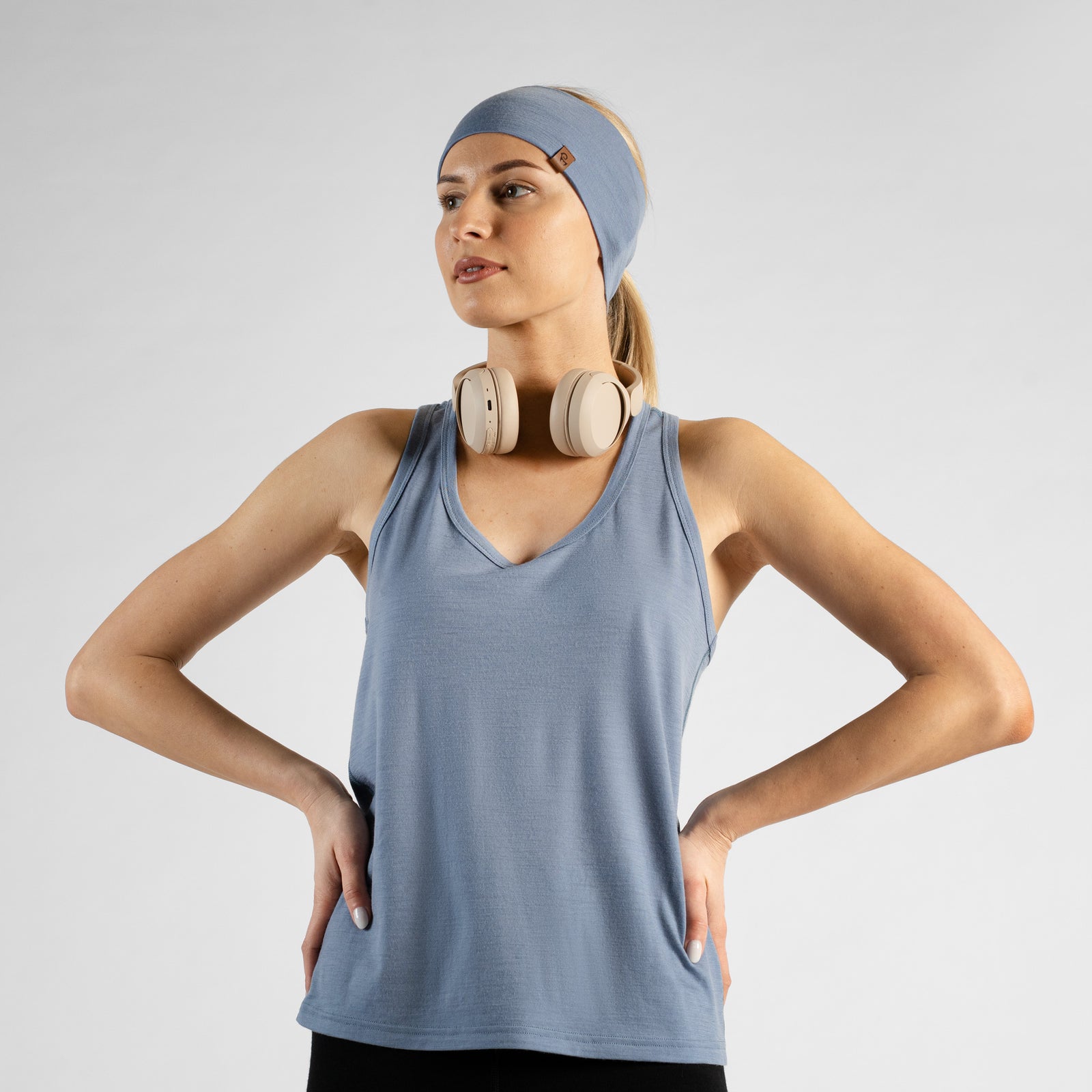 A woman sitting on a white studio floor, viewed from behind. She is wearing a dusty blue racerback tank top, black leggings, and a matching dusty blue headband. Her blonde hair is pulled into a low ponytail, and she has both hands raised behind her head as she adjusts the headband.