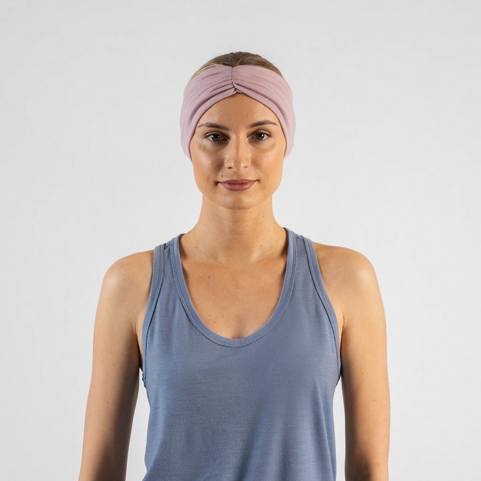A studio portrait of a blonde woman looking slightly off-camera to her left against a plain light grey background. She is wearing a dusty blue sleeveless top and a dusty pink ribbon headband, with her hands gently adjusting the headband near her ears.