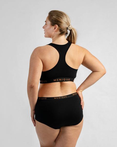 Back view of a model looking over her shoulder with one hand on her hip, wearing a black sports bra and matching boy-short underwear, both featuring a black elastic band with text.