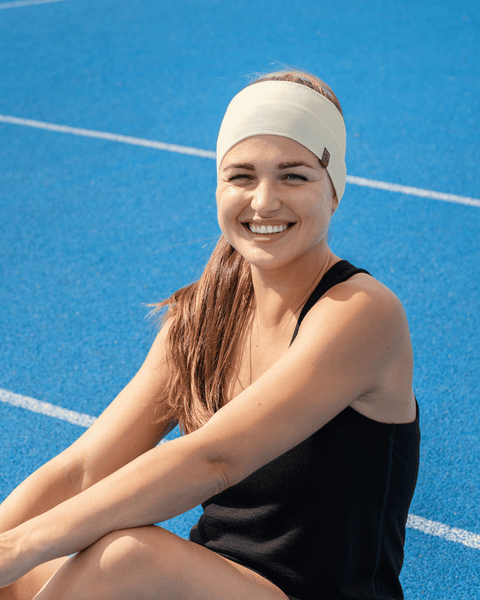 A smiling woman sitting on a blue sports court, wearing a black tank top and a wide white headband with her hands resting on her legs.