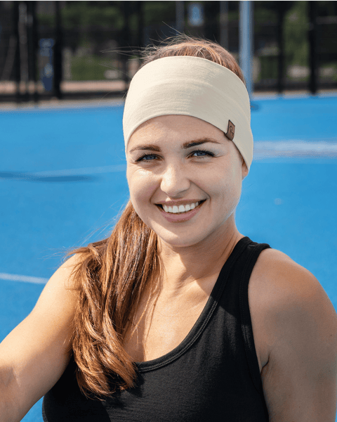 A close-up portrait of a smiling woman outdoors against a blue sports court background, wearing a black tank top and a wide white headband.