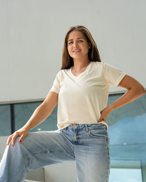 A smiling woman in a cream v-neck t-shirt and blue jeans standing with one leg propped up on a low concrete wall near a glass railing.