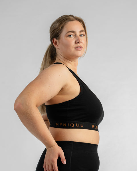 Side profile view of a model looking over her shoulder toward the camera, wearing a black sports bra featuring a black elastic band with text and black bottoms.