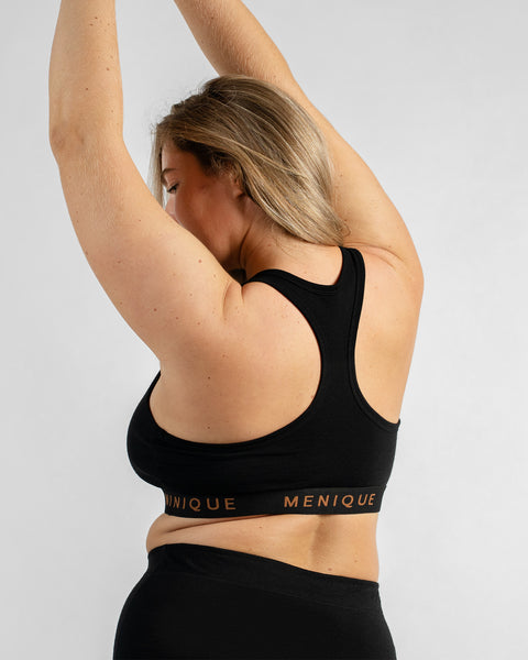 Side-back view of a model with her arms raised, wearing a black sports bra featuring a black elastic band with text and black bottoms.