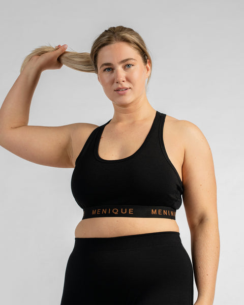 Front view of a model holding her hair up with one hand, wearing a black sports bra featuring a black elastic band with text and black bottoms.
