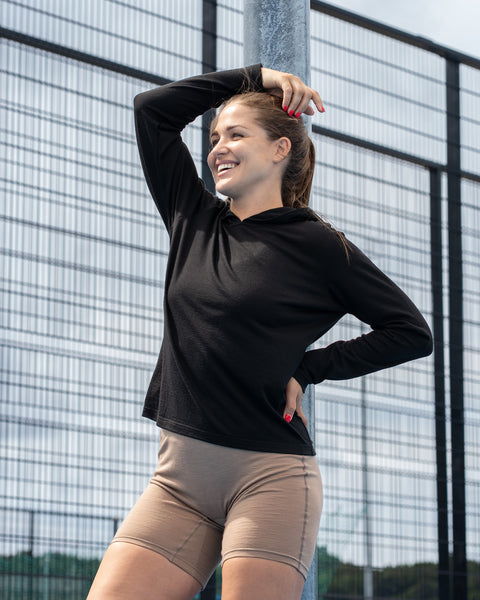 A woman laughing and leaning against a metal pole at an outdoor sports court. She is wearing a black long-sleeve hoodie and tan shorts, with one hand resting near her hair and the other on her hip.