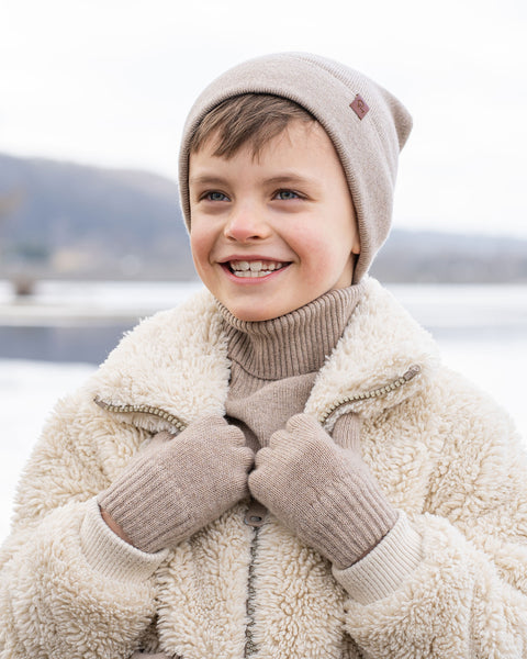 A young child with short brown hair and a wide smile, is dressed in winter clothing. They are wearing a beige beanie, a beige turtleneck sweater, and a cream-colored sherpa jacket, with beige knitted gloves. A snowy, blurred background with what appears to be a body of water is visible behind them.