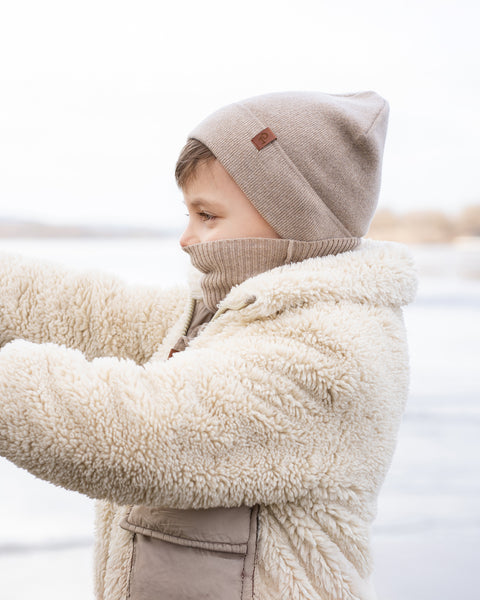 A child with light brown hair, is seen in profile against a blurred, icy landscape. They are wearing a beige beanie, a beige turtleneck, a cream-colored sherpa jacket with pockets, and beige gloves, looking towards the left side of the frame.
