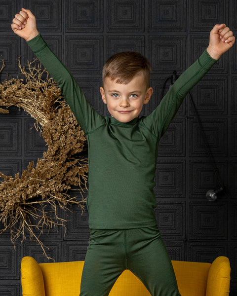 Child in a dark green merino wool outfit with arms raised against a dark textured wall with decorative elements.