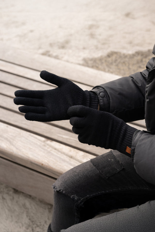 A person wearing black knit gloves from menique, while sitting on a wooden bench outdoors. The gloves appear to be snug-fitting and warm.