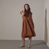 A woman with shoulder-length brown hair models a short-sleeved, almond brown midi linen dress, gently swaying and posing in a brightly lit corner of a room in a brief video.